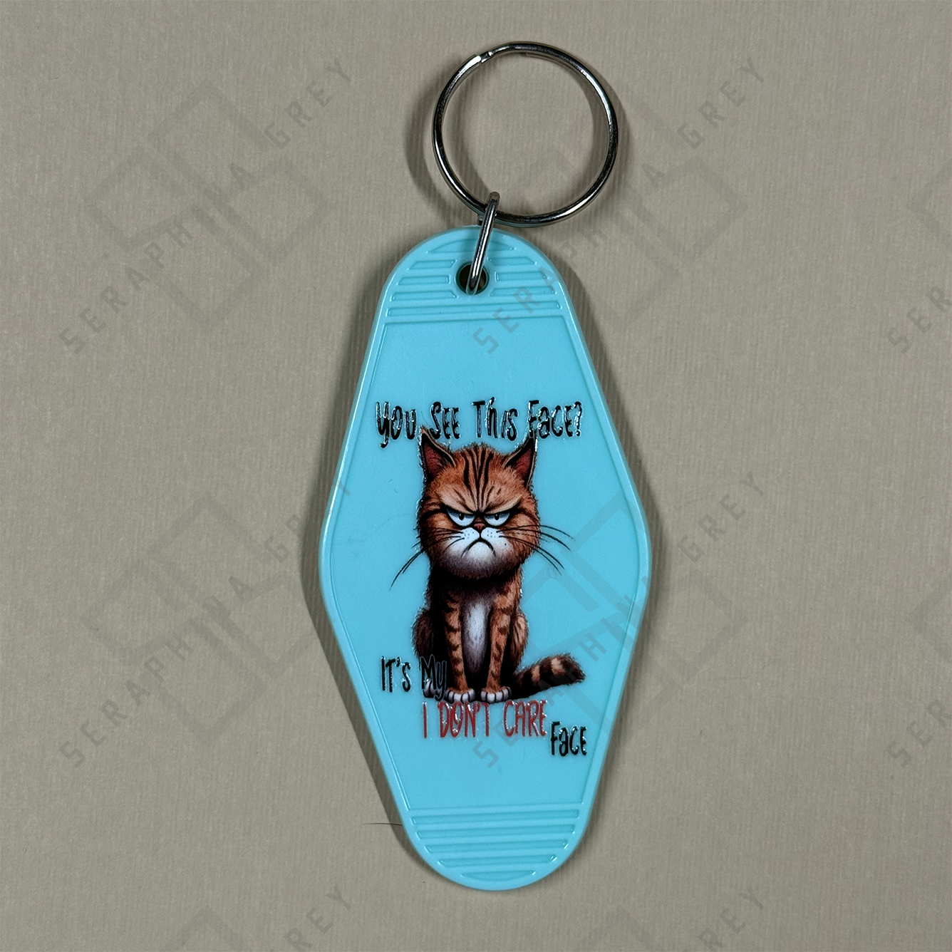 Blue keychain with a grumpy cat illustration and text  you see this face, its my I dont care face. on a beige background